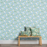 A stool stacked with throw pillows in front of a wall papered in a floral print in white, green and blue.