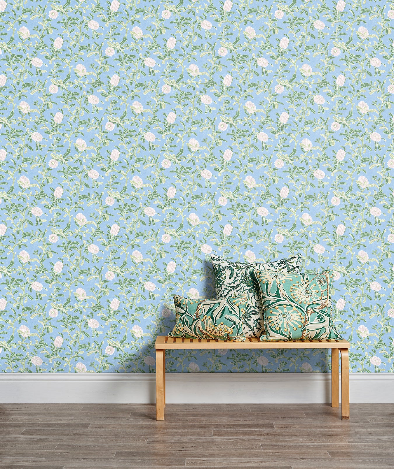 A stool stacked with throw pillows in front of a wall papered in a floral print in white, green and blue.