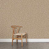 A wooden chair stands in front of a wall papered in a minimal repeating leaf print in cream on a light brown field.