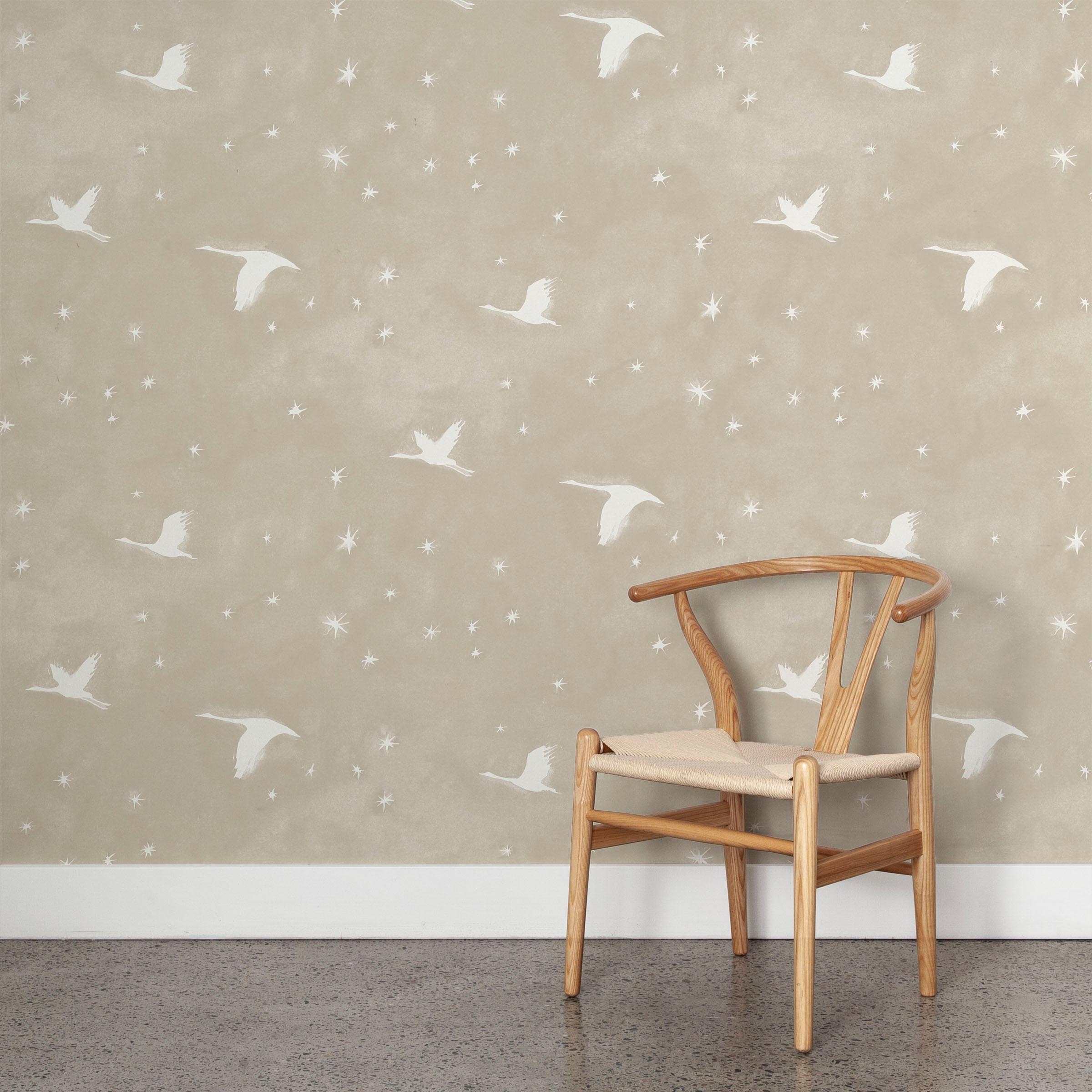 A wooden chair stands in front of a wall papered in a print of birds and stars in white on a tan watercolor field.