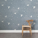 A wooden chair stands in front of a wall papered in a print of birds and stars in white on a light blue watercolor field.