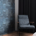 A patterned armchair stands in front of a wall papered in square leather tiles in mottled navy.