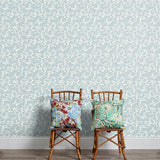 Two wooden chairs stand in front of a wall papered in a dense leaf and stem print in blue-gray and white.