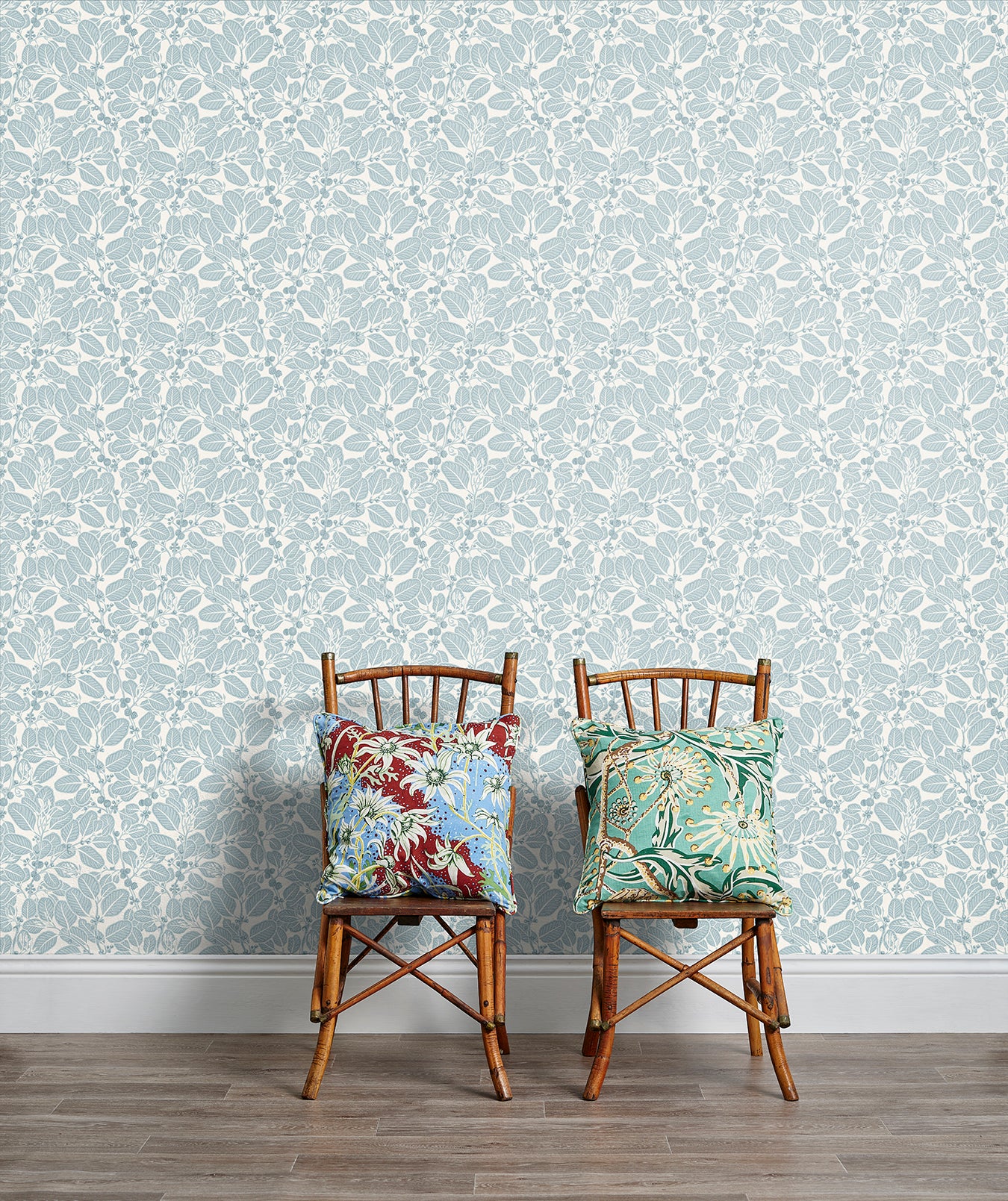 Two wooden chairs stand in front of a wall papered in a dense leaf and stem print in blue-gray and white.