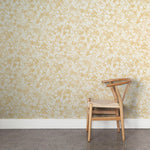 A wooden chair stands in front of a wall papered in a painterly branch print in white on a yellow watercolor field.