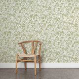 A wooden chair stands in front of a wall papered in a painterly branch print in white on a light green watercolor field.
