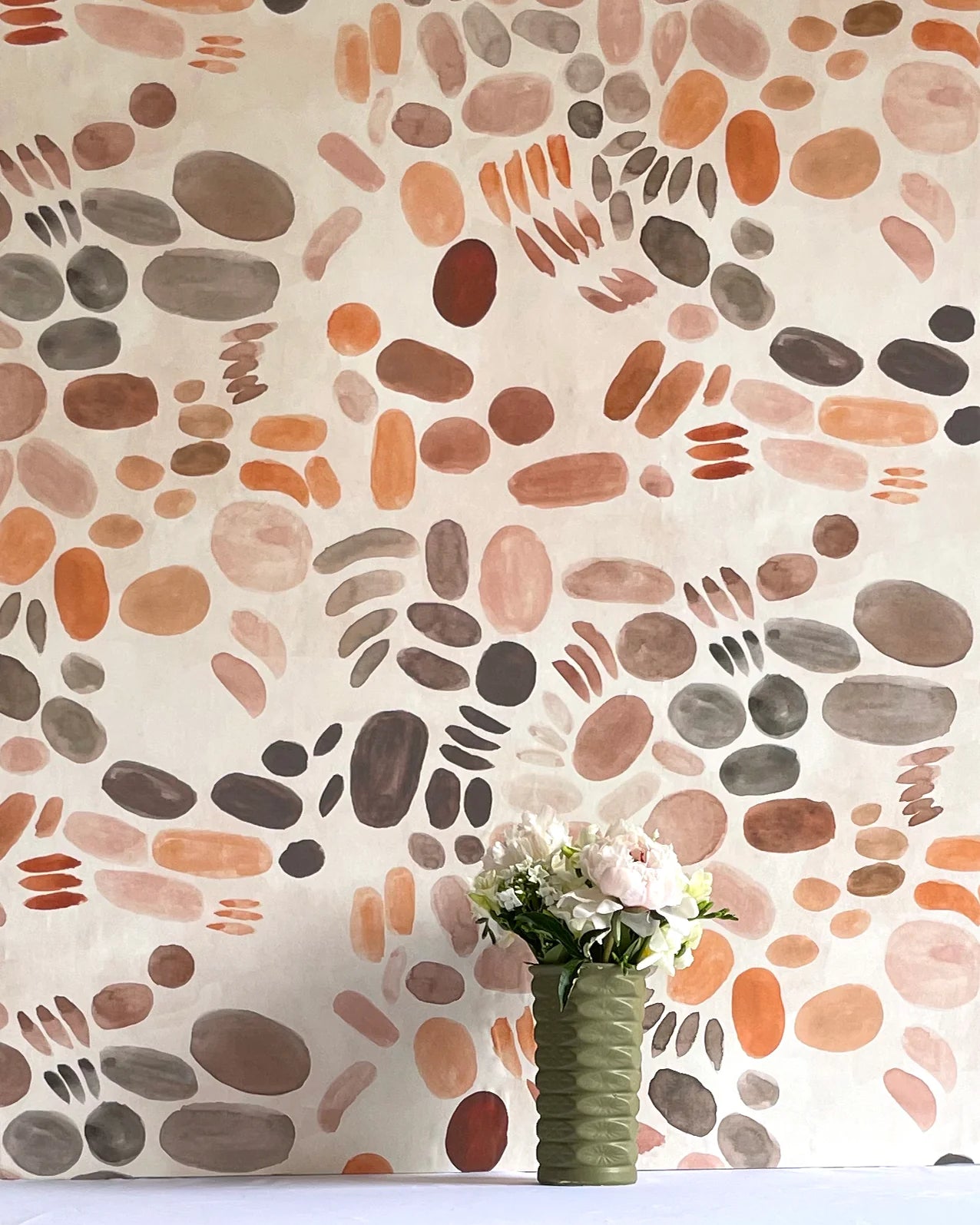 A vase of flowers stands in front of a wall covered in a playful paint blotch print in brown and orange on a cream field.