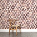 A wooden chair stands in front of a wall papered in a painterly small-scale diamond print in cream, brown, purple and pink.