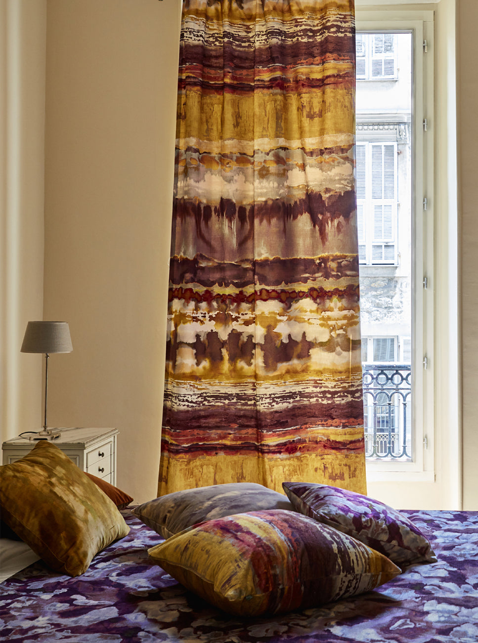 A sunny bedroom with curtains and throw pillows all in the same abstract paint swatch print.