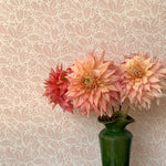 A green vase with pink peonies against a botanical leaf patterned wallpaper in soft pink.