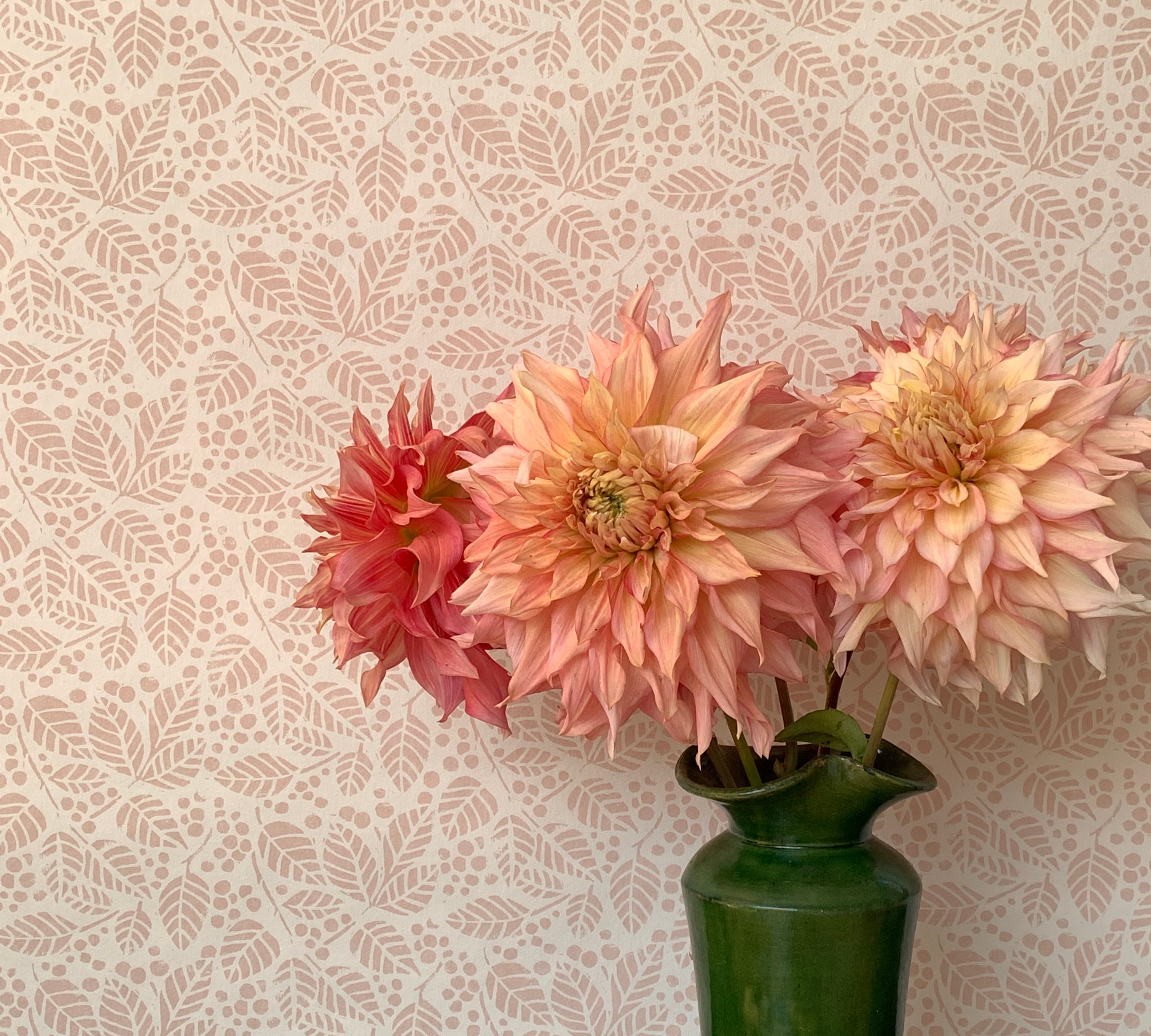 A green vase with pink peonies against a botanical leaf patterned wallpaper in soft pink.