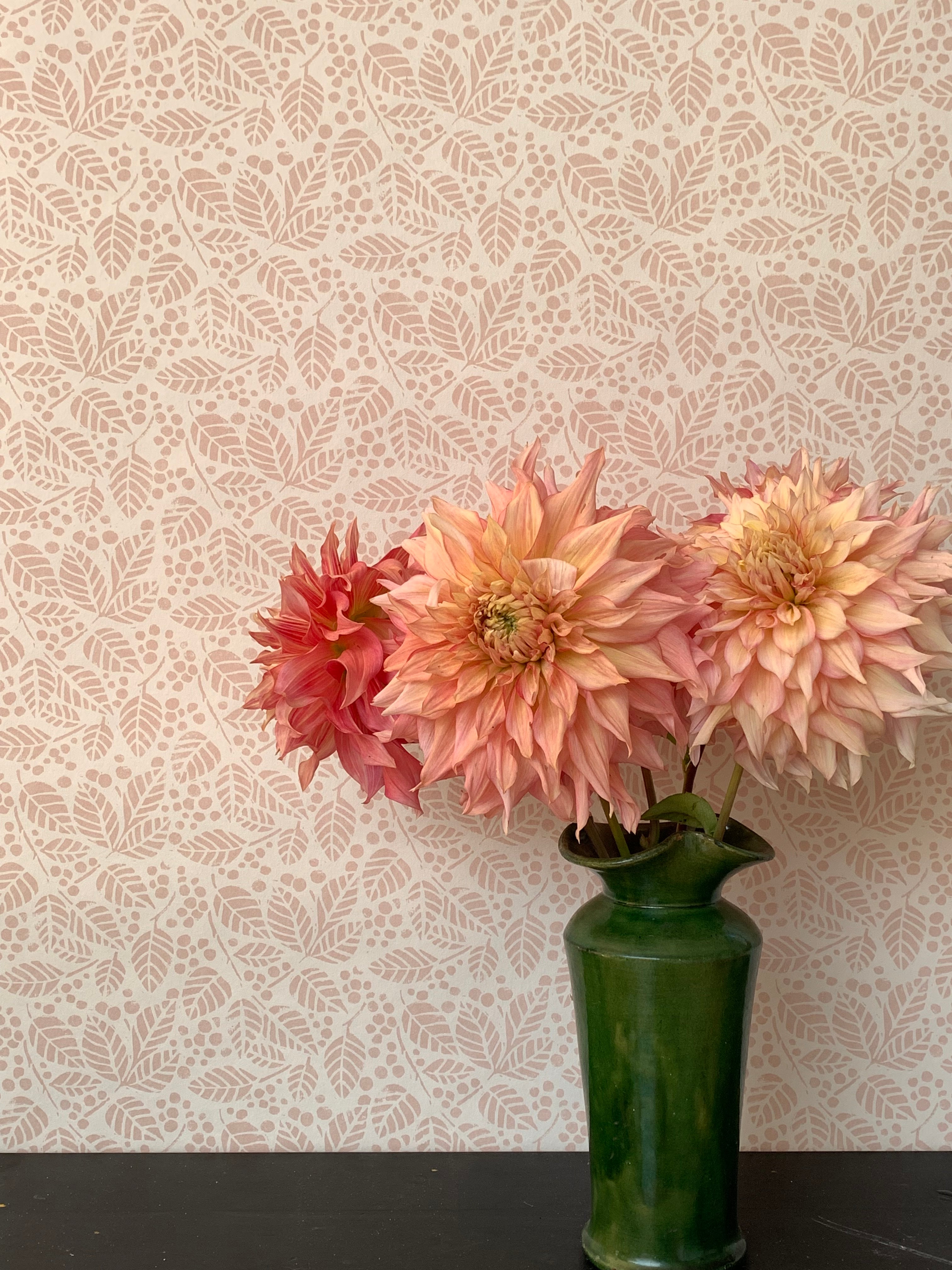 A green vase with pink peonies against a botanical leaf patterned wallpaper in soft pink.