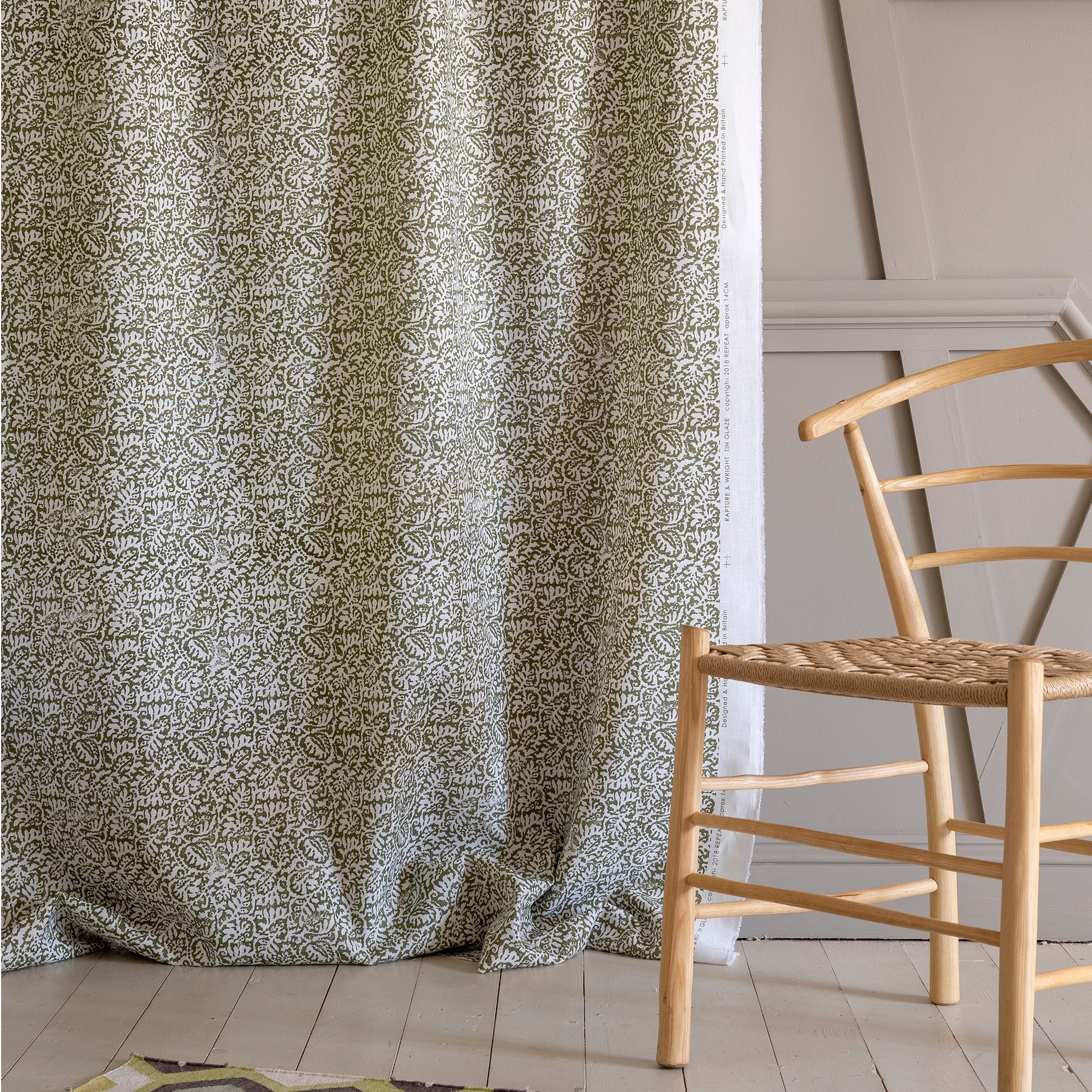 A wooden chair stands in front of draped fabric yardage in a dense paisley print in white on a mottled green field.