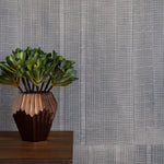 A potted plant stands in front of a wall papered in an irregular grid pattern in charcoal on a gray field.