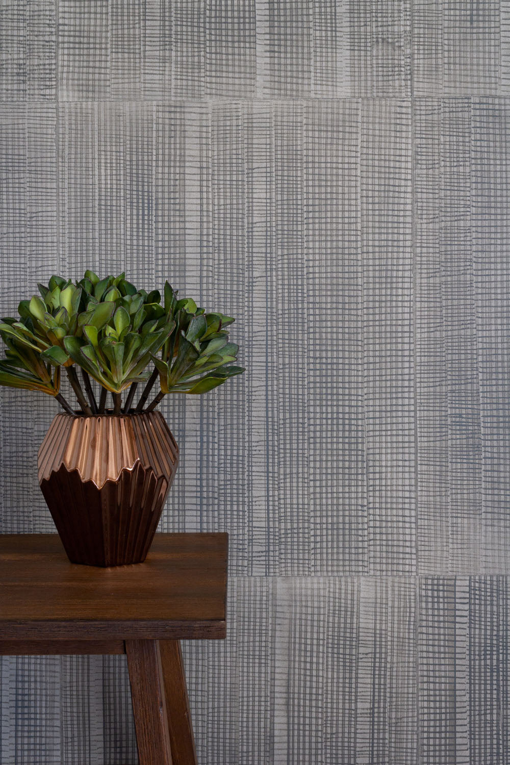 A potted plant stands in front of a wall papered in an irregular grid pattern in charcoal on a gray field.