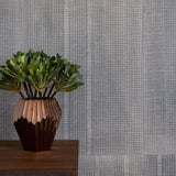 A potted plant stands in front of a wall papered in an irregular grid pattern in charcoal on a gray field.