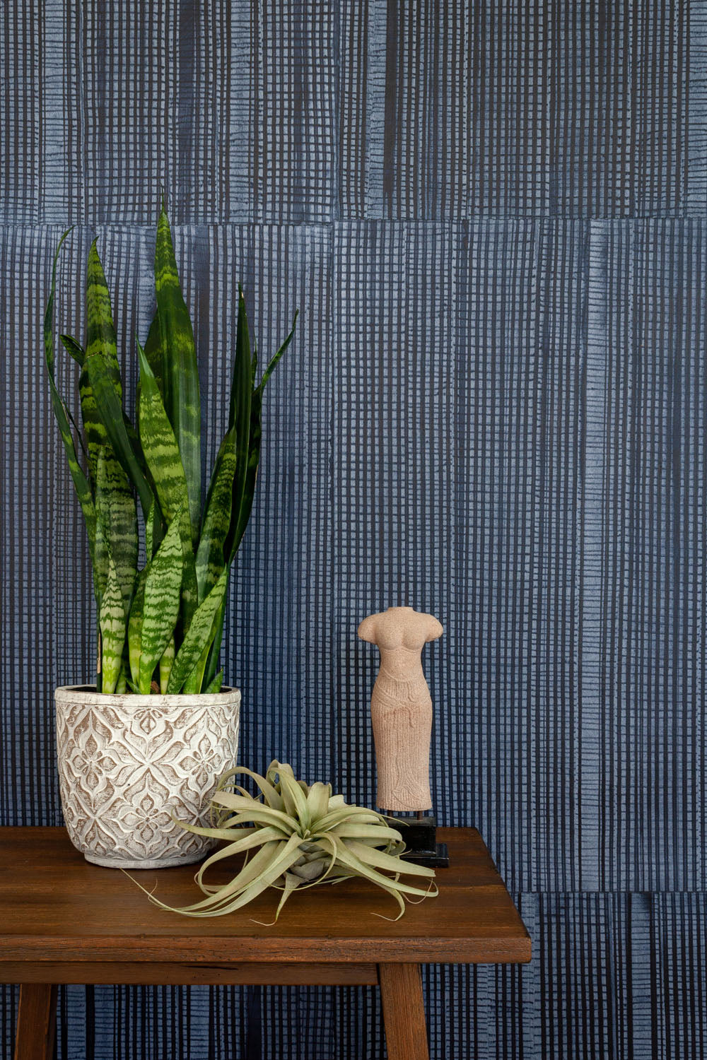 Plants and a statue stand in front of a wall papered in an irregular grid pattern in charcoal on a navy field.