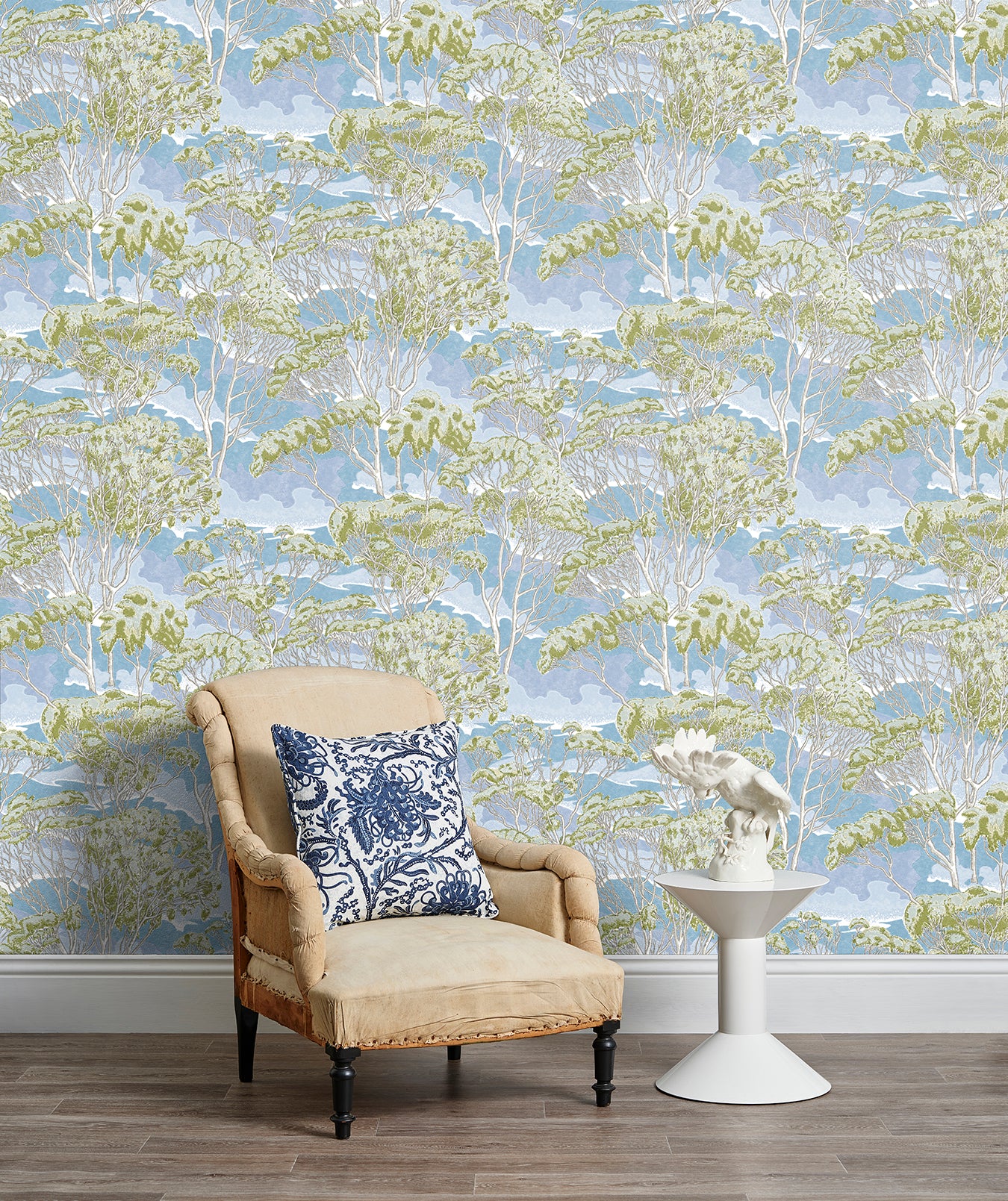 Armchair and end table in front of a wall papered in a painterly tree print in cream, green and light blue.