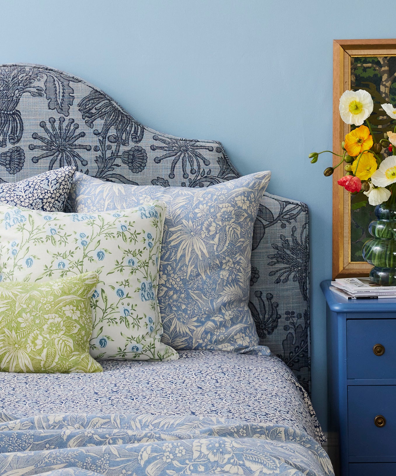 Bed with floral bedding and pillows against a blue wall, next to a blue nightstand with decorative items.