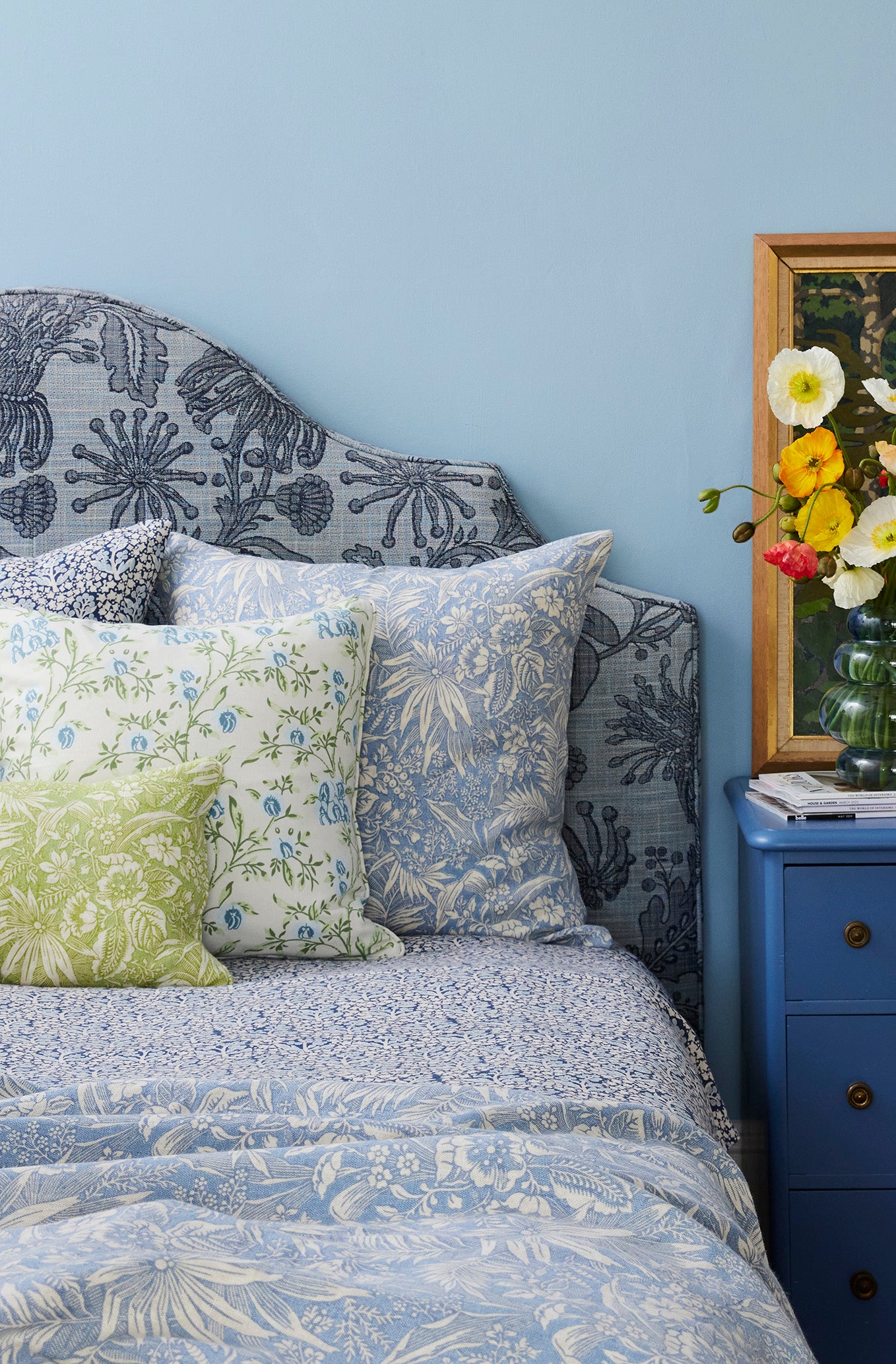 Bed with floral bedding and pillows against a blue wall, next to a blue nightstand with decorative items.