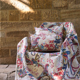 Chair and pillows in a  lyrical jungle design with tropical birds and leafy branches in pink, tan, blue and yellow against a tan stone wall. 