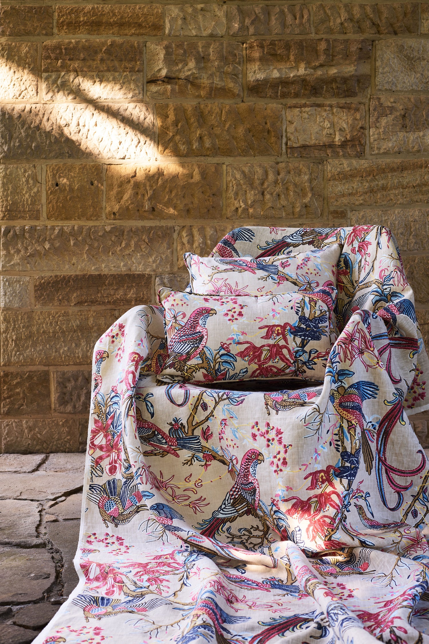 Chair and pillows in a  lyrical jungle design with tropical birds and leafy branches in pink, tan, blue and yellow against a tan stone wall. 