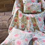 Pillows atop a fabric draped chair all in a matching printed fabric in a lush floral botanical design in red, pink, yellow green and turquoise on a tan field against a stone wall. 