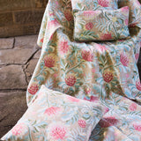 Pillows atop a fabric draped chair all in a matching printed fabric in a lush floral botanical design in red, pink, yellow green and turquoise on a tan field against a stone wall. 