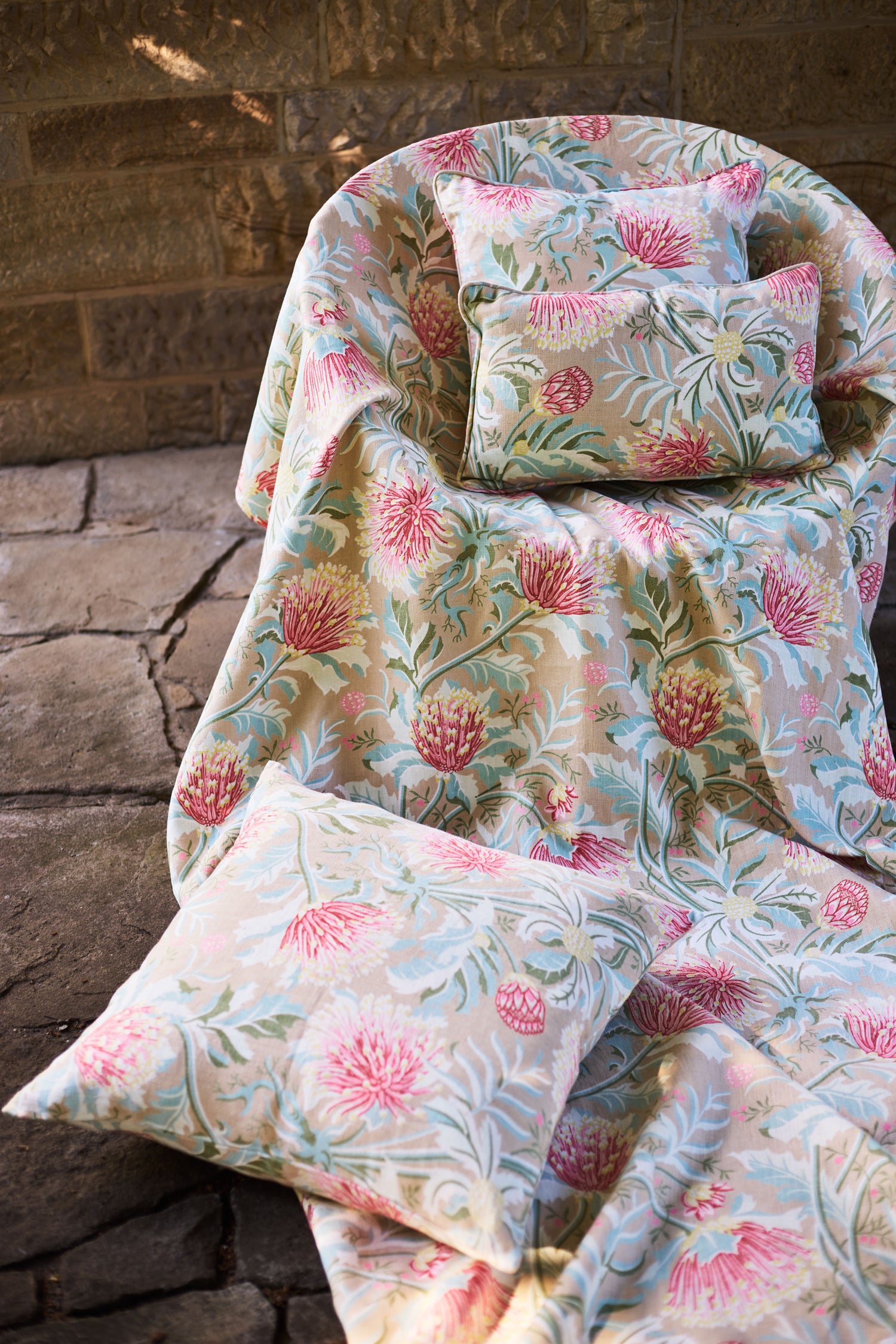 Pillows atop a fabric draped chair all in a matching printed fabric in a lush floral botanical design in red, pink, yellow green and turquoise on a tan field against a stone wall. 