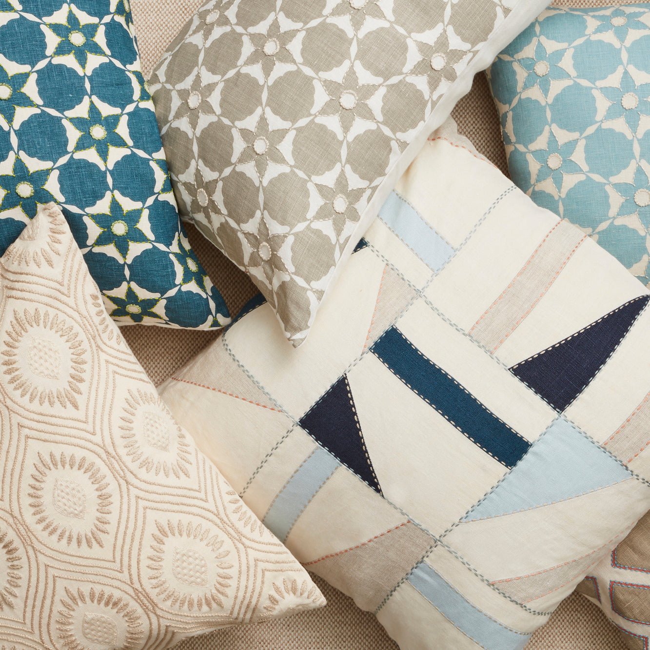 A pile of throw pillows on a tan surface. Throw pillows are patterned in shades of cream, light blue and navy.