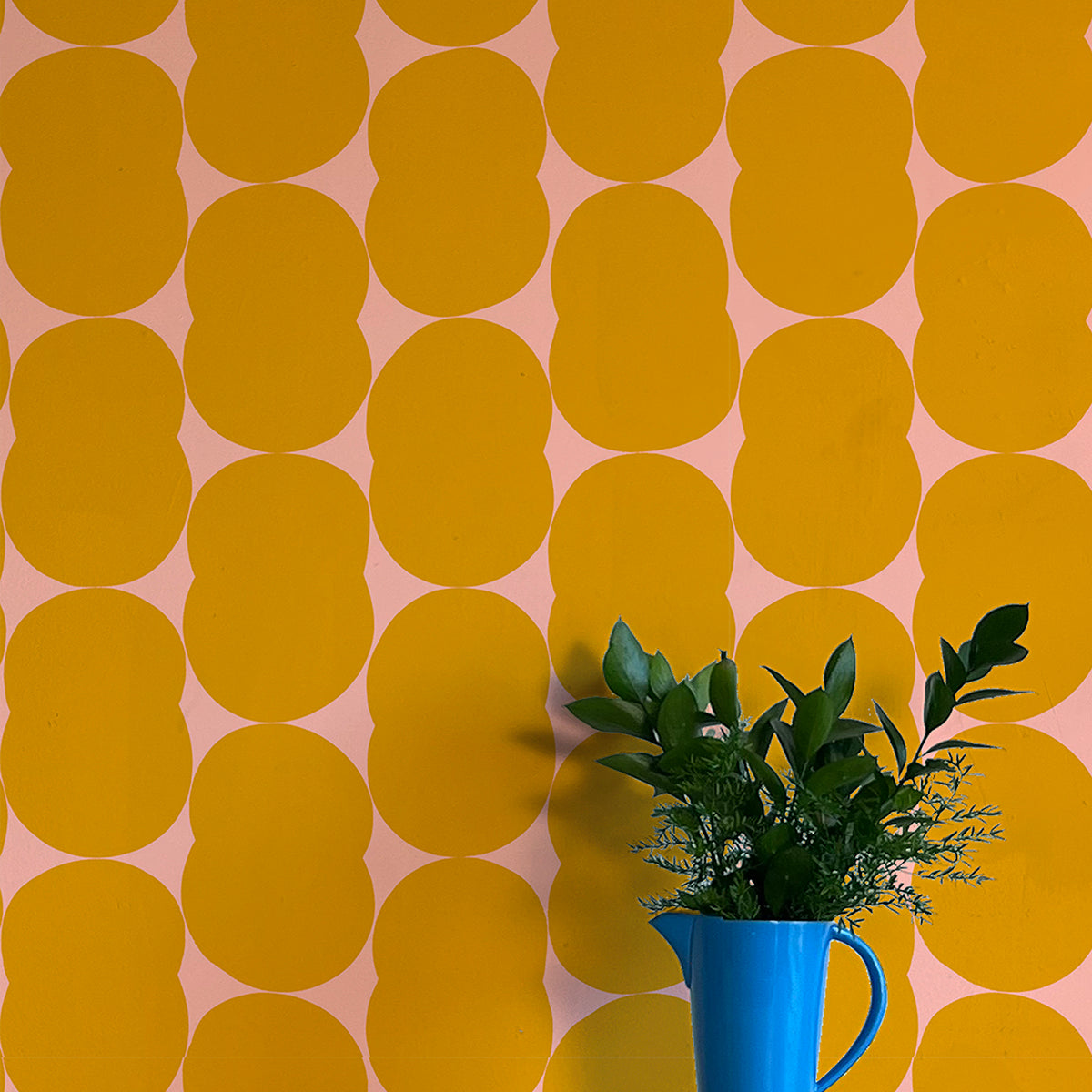 A vase of flowers stands in front of a wall papered in an abstract curvilinear pattern in mustard on a pink field.