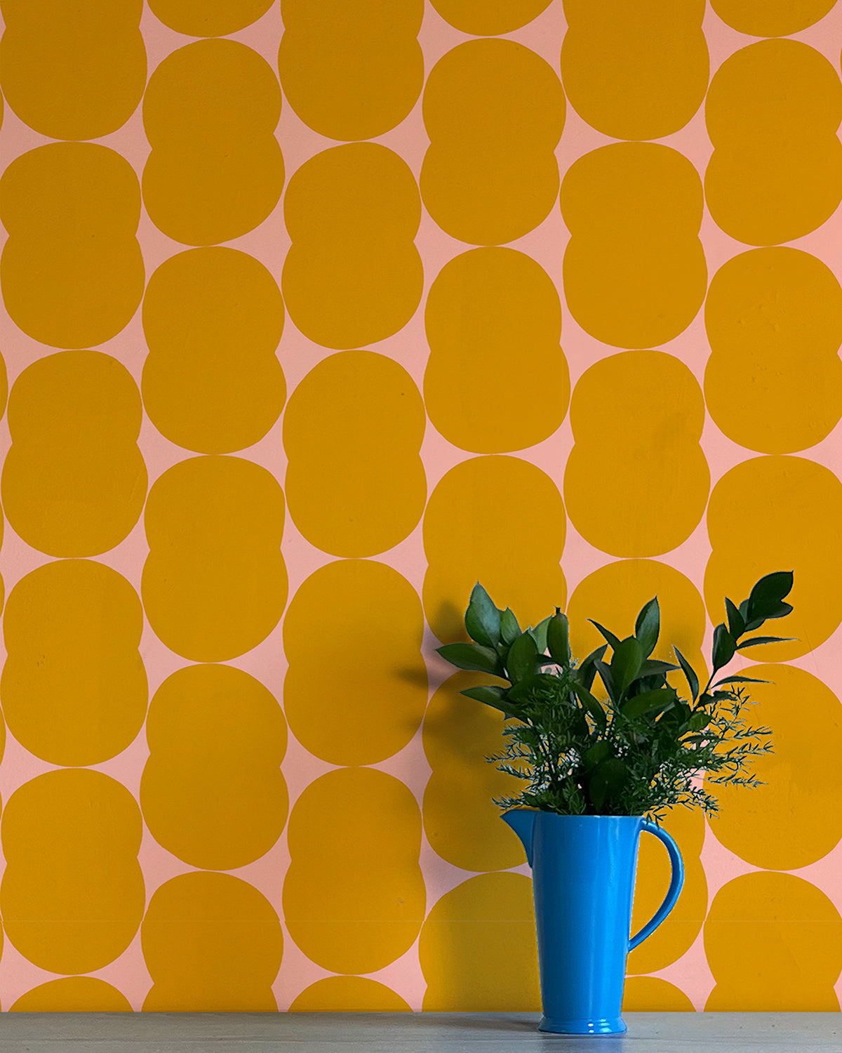 A vase of flowers stands in front of a wall papered in an abstract curvilinear pattern in mustard on a pink field.