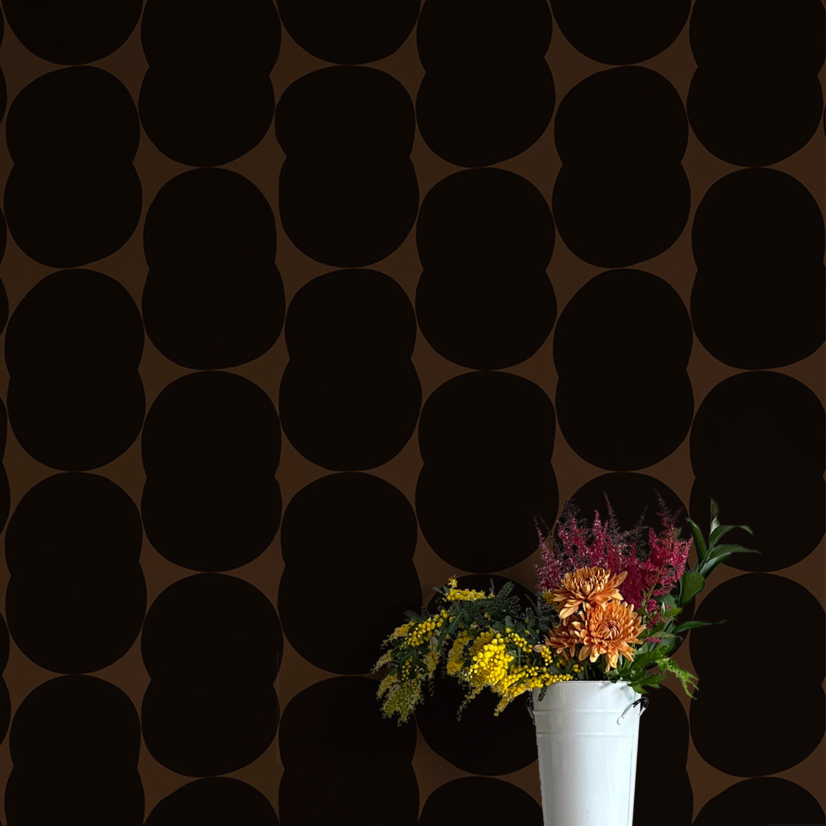 A vase of flowers stands in front of a wall papered in an abstract curvilinear pattern in black on a brown field.