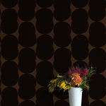 A vase of flowers stands in front of a wall papered in an abstract curvilinear pattern in black on a brown field.