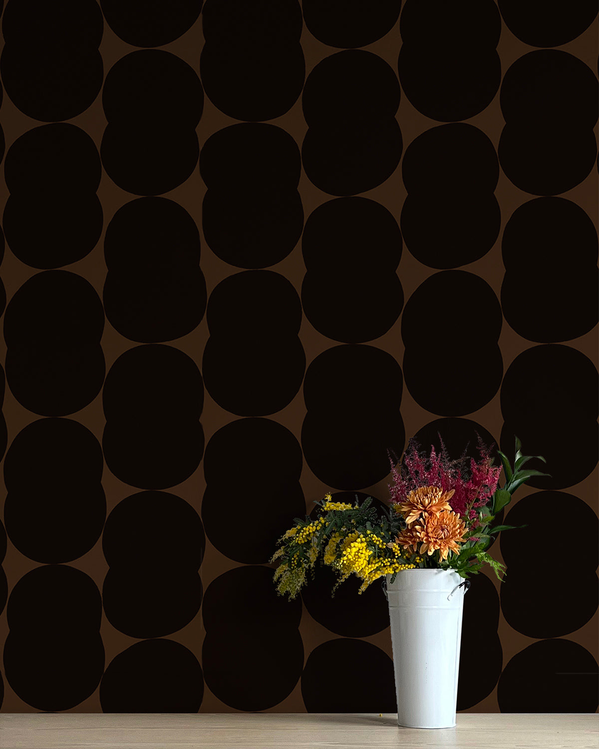 A vase of flowers stands in front of a wall papered in an abstract curvilinear pattern in black on a brown field.