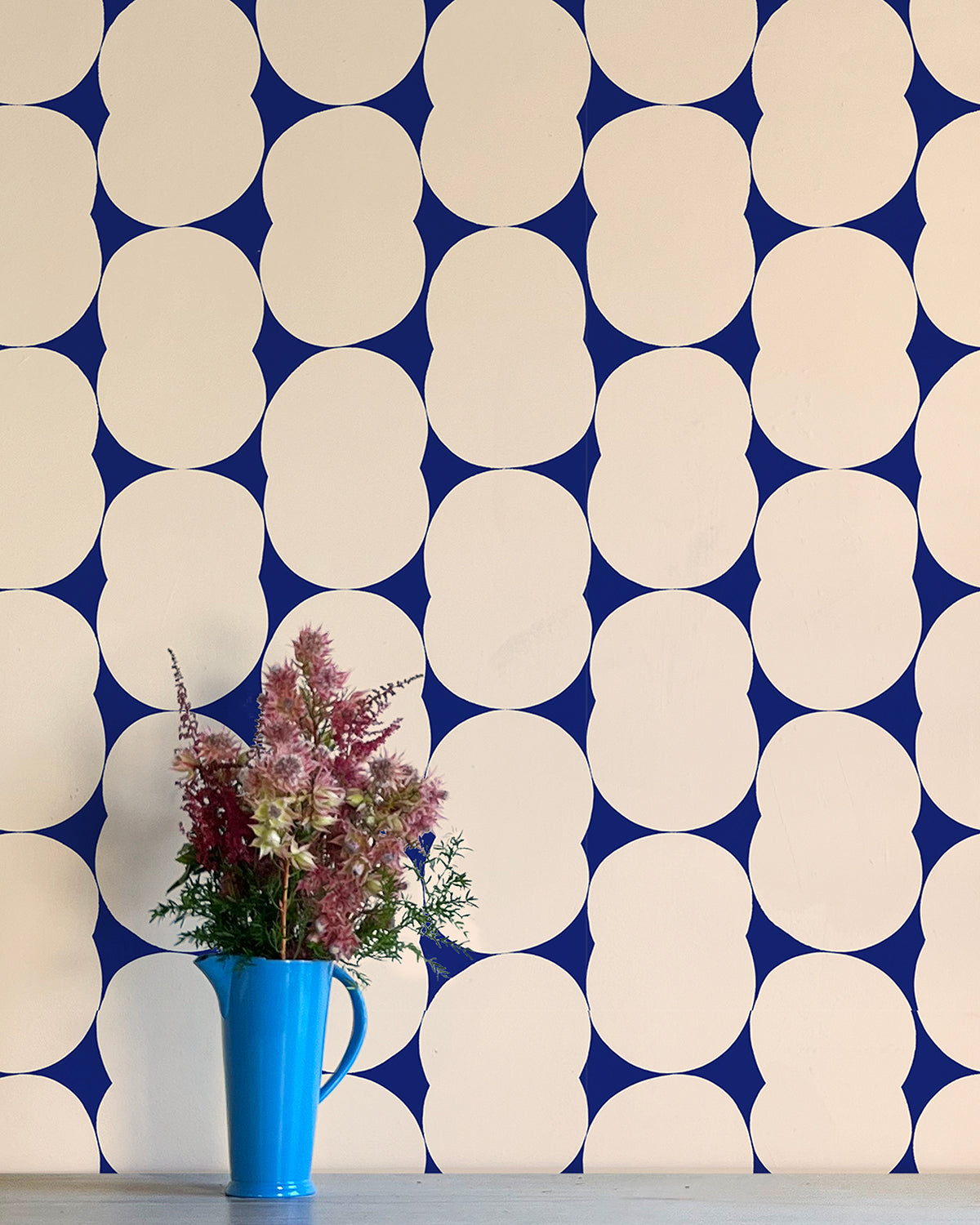 A vase of flowers stands in front of a wall papered in an abstract curvilinear pattern in cream on a navy field.