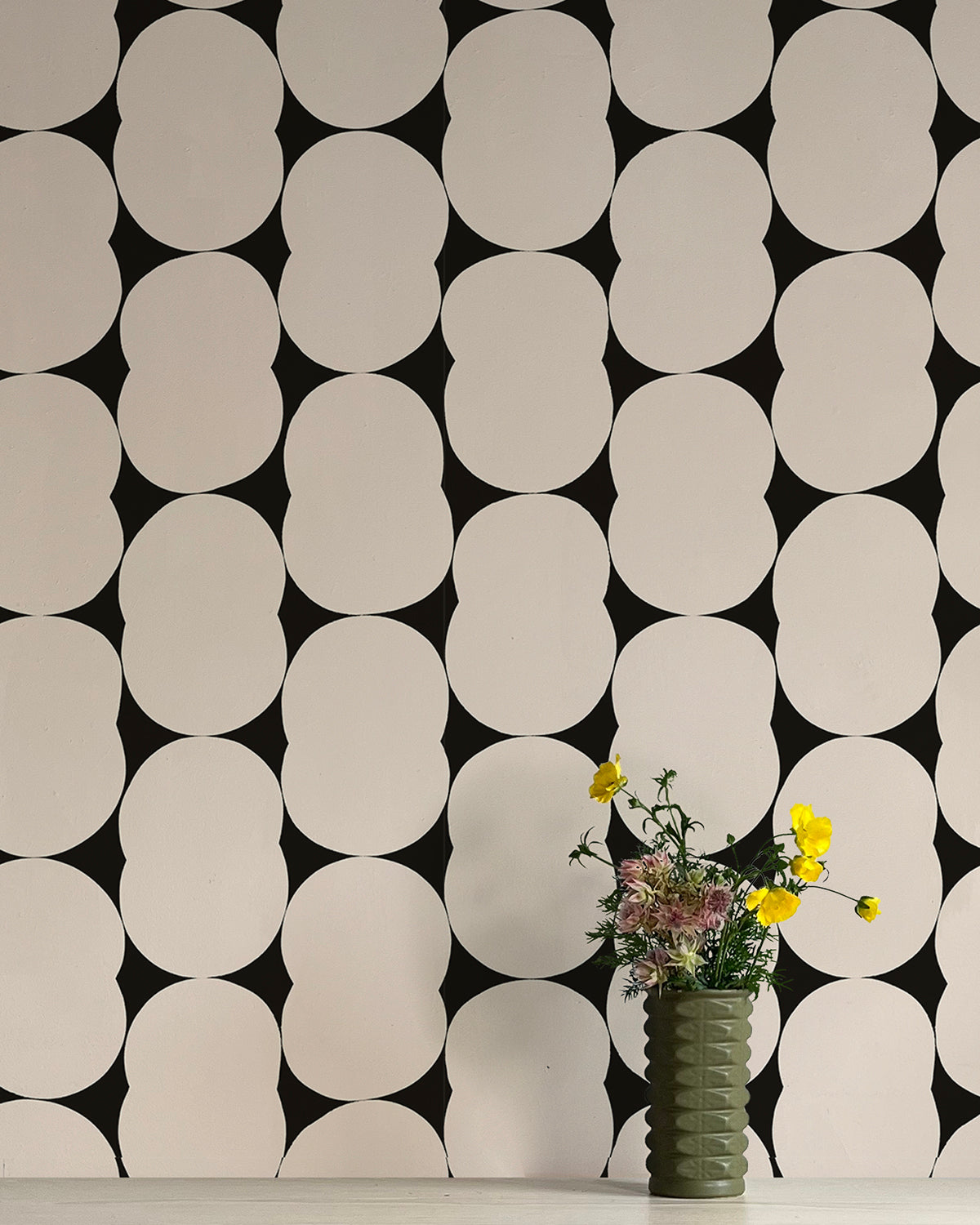 A vase of flowers stands in front of a wall papered in an abstract curvilinear pattern in gray on a black field.