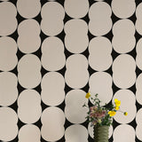 A vase of flowers stands in front of a wall papered in an abstract curvilinear pattern in gray on a black field.