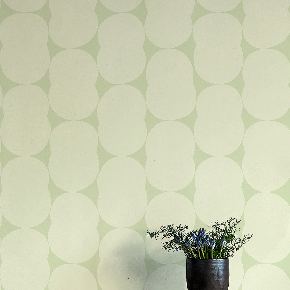 A vase of flowers stands in front of a wall papered in an abstract curvilinear pattern in cream on a light green field.