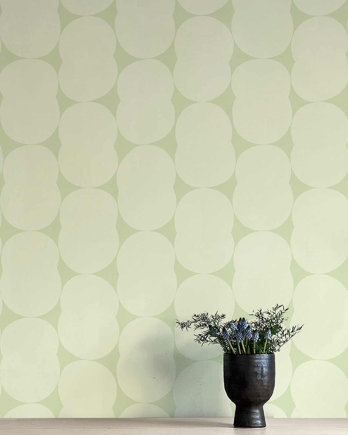 A vase of flowers stands in front of a wall papered in an abstract curvilinear pattern in cream on a light green field.