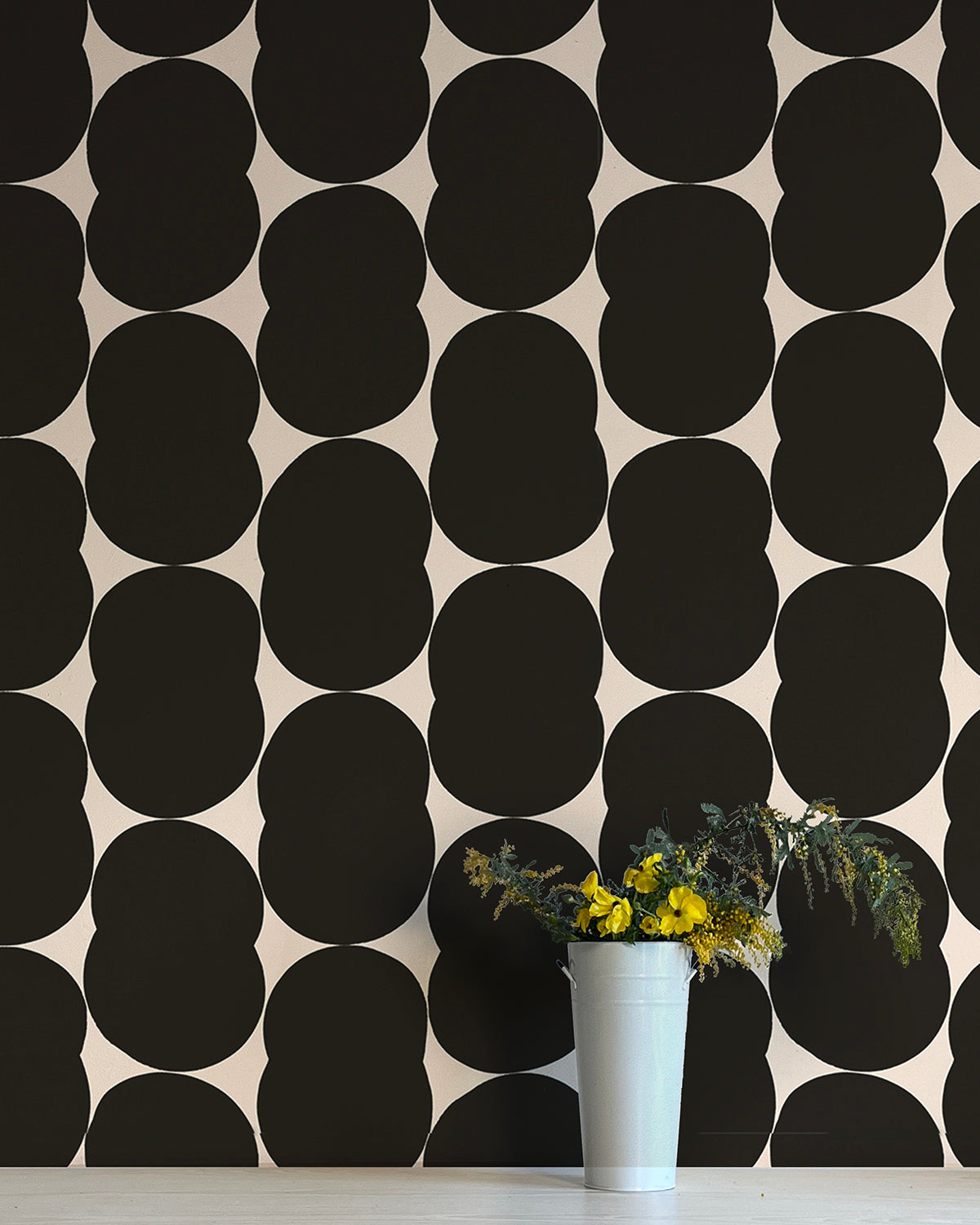 A vase of flowers stands in front of a wall papered in an abstract curvilinear pattern in black on a white field.