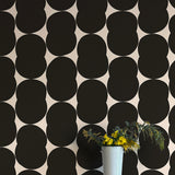 A vase of flowers stands in front of a wall papered in an abstract curvilinear pattern in black on a white field.