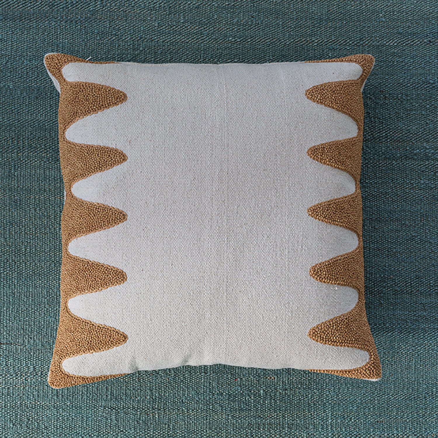 Square cream throw pillow with a wavy raffia embroidery pattern on the sides, laying on a turquoise fabric background.