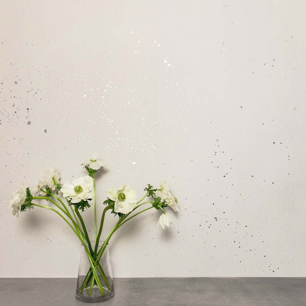 A vase of white flowers stands in front of a wall papered in a random splattered pattern in metallic gold on a cream field.