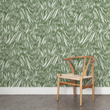 A wooden chair stands in front of a wall papered in an abstract leaf pattern in white on a sage watercolor field.