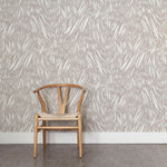 A wooden chair stands in front of a wall papered in an abstract leaf pattern in white on a light gray watercolor field.