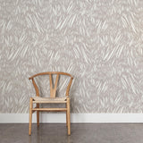 A wooden chair stands in front of a wall papered in an abstract leaf pattern in white on a light gray watercolor field.