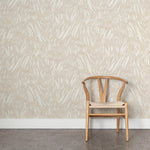 A wooden chair stands in front of a wall papered in an abstract leaf pattern in white on a cream watercolor field.
