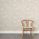 A wooden chair stands in front of a wall papered in an abstract leaf pattern in white on a cream watercolor field.