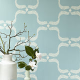 An end table with flowers stands in front of a wall papered in a leafy lattice print in cream on a light blue field.